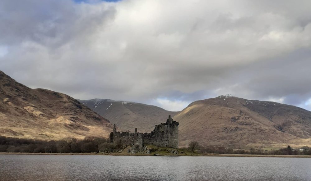 Old castle ruin by a lake with mountains in the background under cloudy sky.