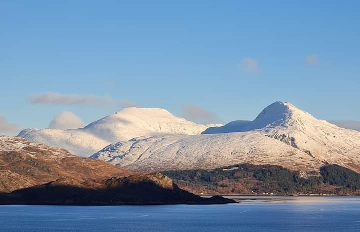 Snow-covered mountains under a clear blue sky above calm water.